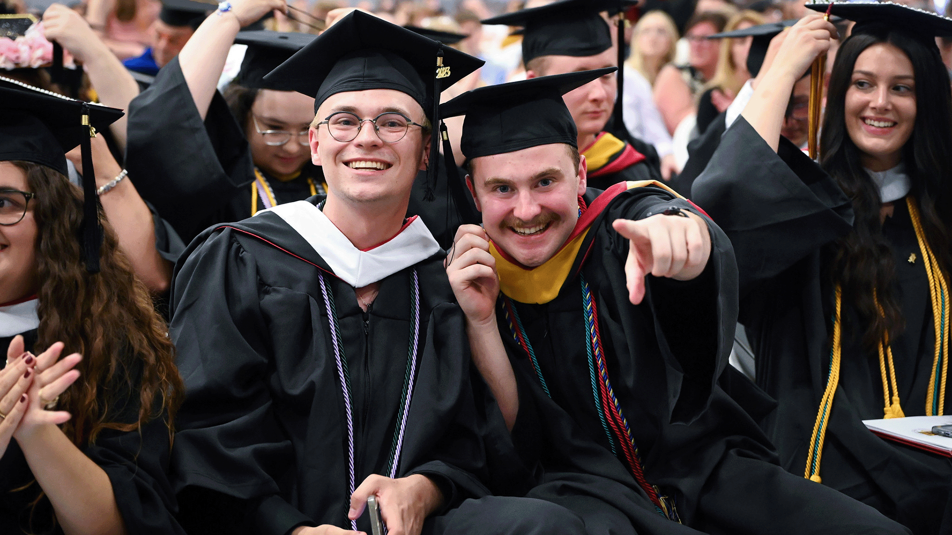 Students in commencement regalia with one graduate pointing at the viewer.