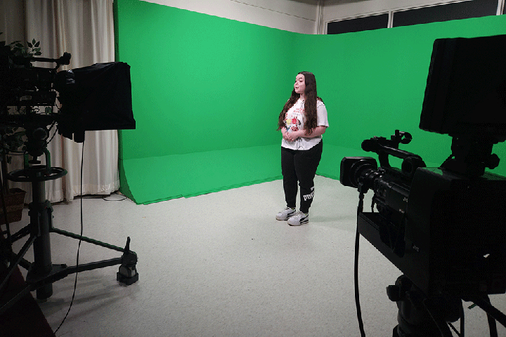 A student stands in front of a green screen on camera.