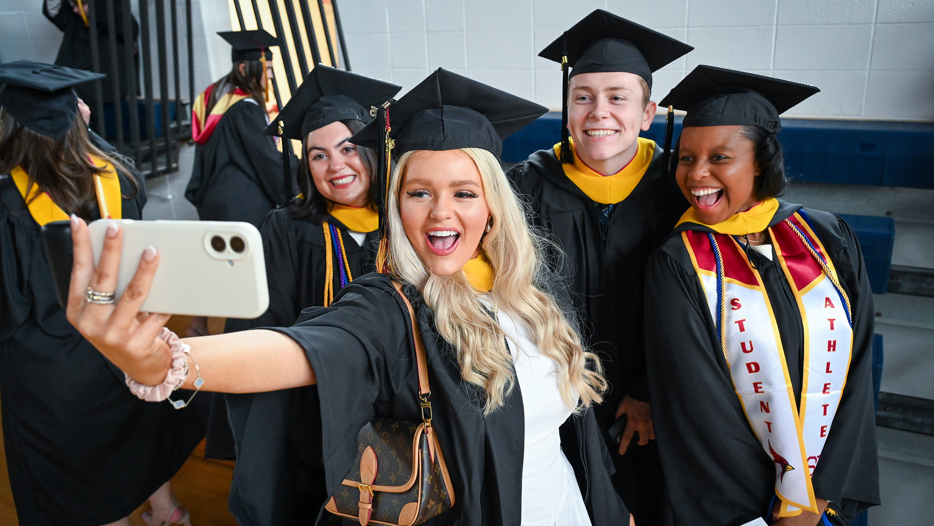 A graduating student takes a selfie with a group dressed in commencement regalia.