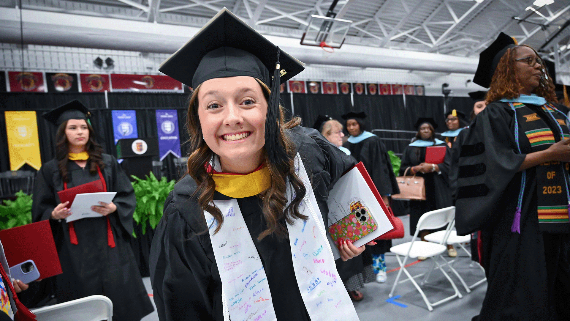 A student in commencement regalia leans forward with a huge smile.