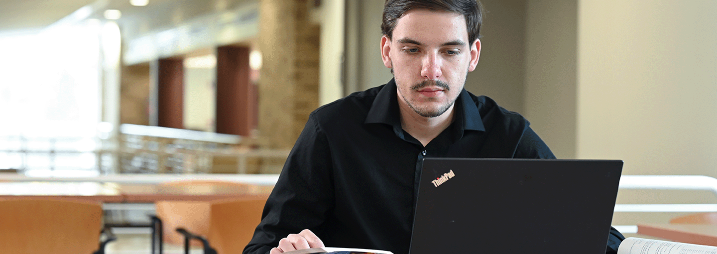 A student studies at a laptop with an open book.