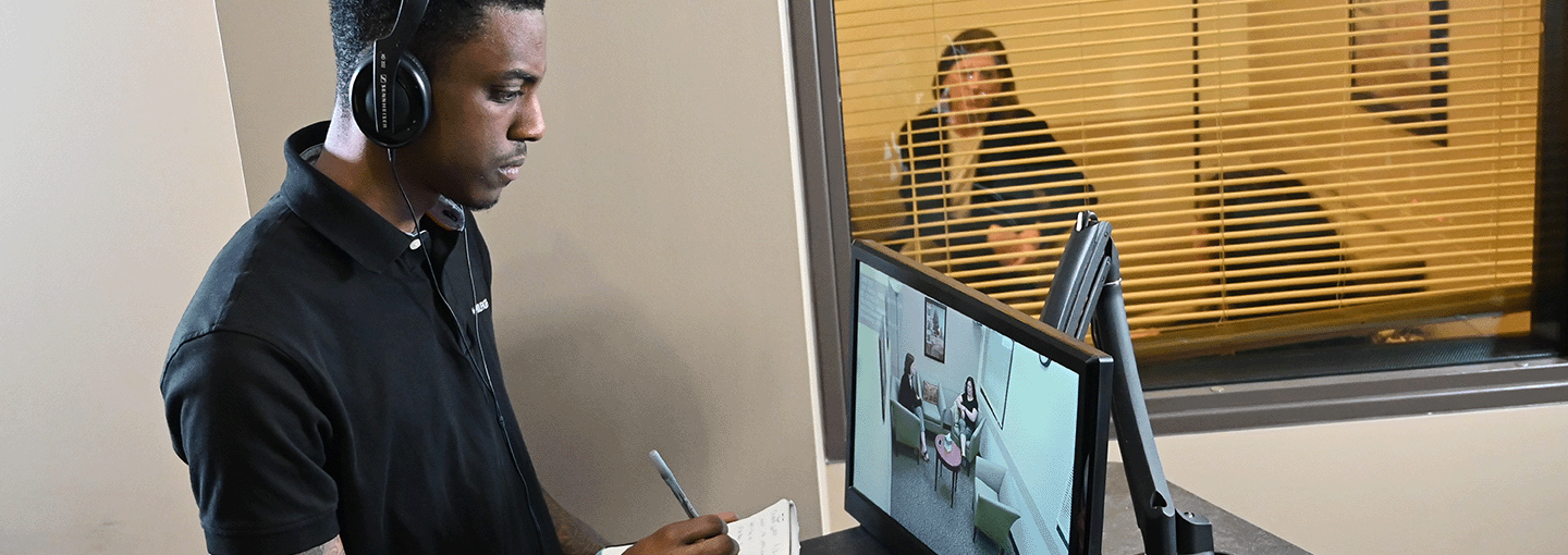 A mental health counseling student writes on a notepad while viewing a counseling simulation.